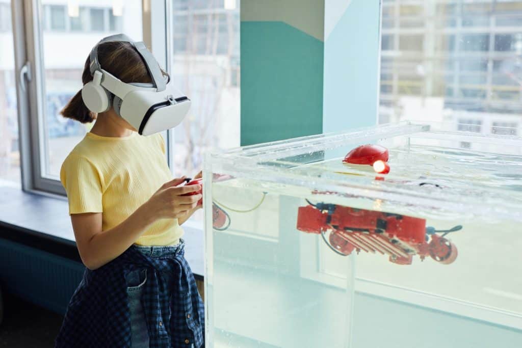 Side view of little girl wearing VR goggles and exploring new robot with controllers in light room